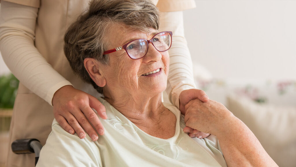 Eine ältere Dame sitz lächelnd im Rollstuhl und blickt aus dem Fenster, während ihre Betreuerin hinter ihr steht und sich an der Hand halten.