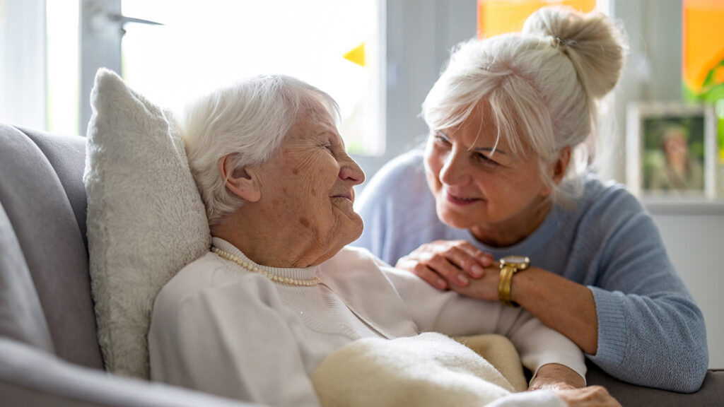 Ihre 24-Stunden-Betreuung.Eine ältere Frau liegt im Bett und unterhält freundlich sich mit ihrer Pflegerin.