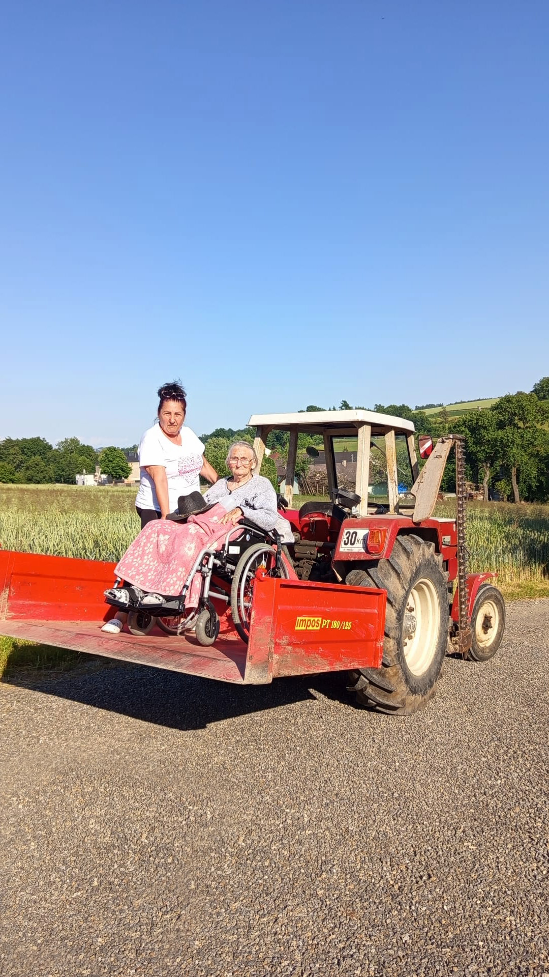 Über uns 9 Eine ältere Dame im Rollstuhl und ihre Betreuerin stehen hinten auf der Ladefläche von einem Traktor. Sie haben den Nachbarn einen Besuch abgestattet.