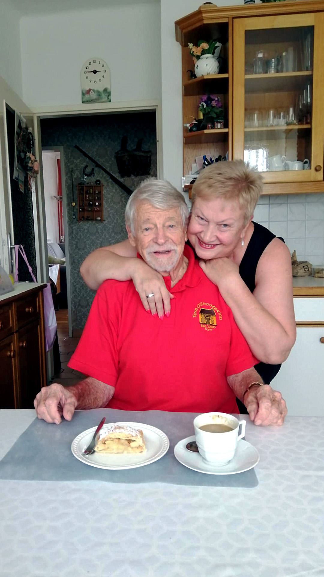Über uns 4 Pflegerin von Lumen 24Pflege umarmt einen älteren Herren. Beide lächeln freundlich in die Kamera. Der Herr sitzt beim Esstisch. Dieser ist gedeckt mit einem Apfelstrudel und Kaffee.