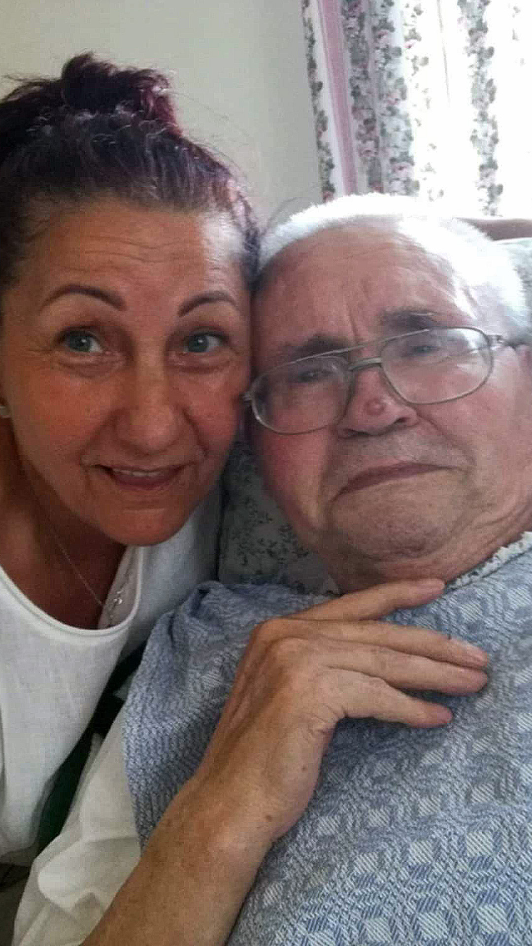 Über uns 8 Älterer Herr liegt im Bett und seine Betreuerin steht links neben ihm und umarmt ihn. Beide lächeln dabei herzlich in die Kamera.